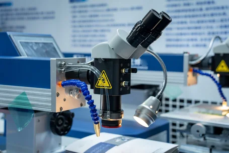 Testa di lavorazione laser che salda un componente in plastica trasparente con un fascio laser rosso, monitor che mostrano il cordone di saldatura a fianco – saldatura laser IR e trasmissione