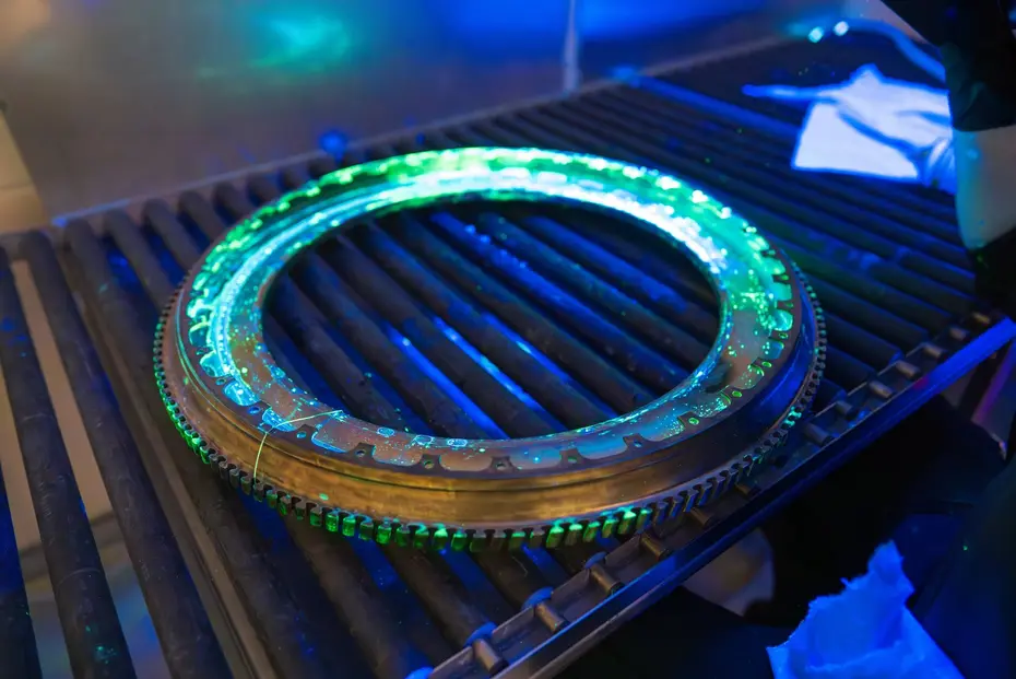 Ring-shaped, toothed metal component in a darkened inspection booth with green fluorescent penetrant indications along the tooth flanks under UV-A illumination, inspector with cloth in the background