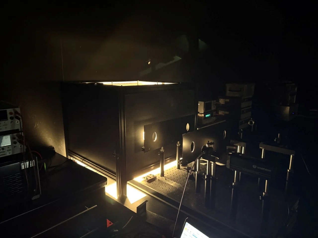 Kalibratie in het verduisterde laboratorium: een FEL-standaardlamp in de zwartgemaakte behuizing bestraalt via twee diafragma's op de optische bank het testobject; links de voedingsbron van de lamp